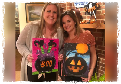 Two women holding up halloween paintings in front of a fireplace.